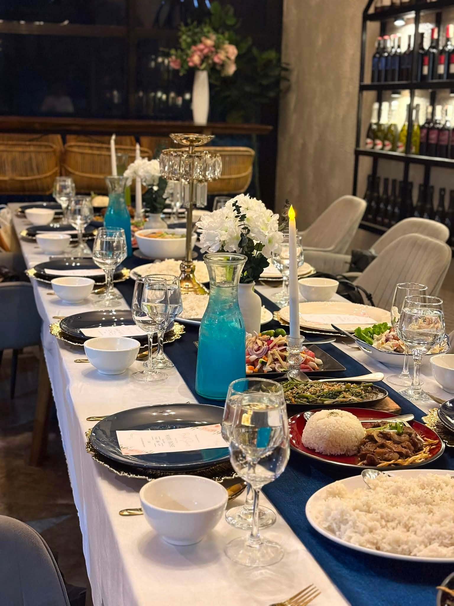 A long dinner table with rice, beef, sautéed greens and salads beside the wine shelves