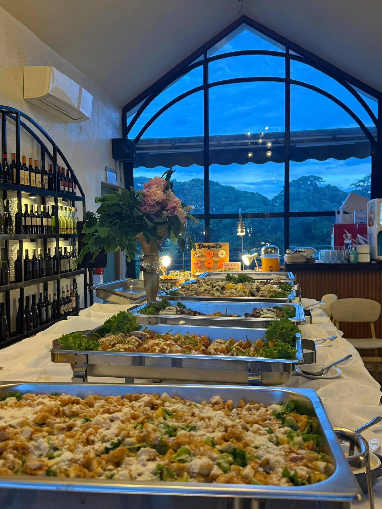 A buffet laid out beneath the tall arched window at dusk, with hills outside and the wine shelves alongside