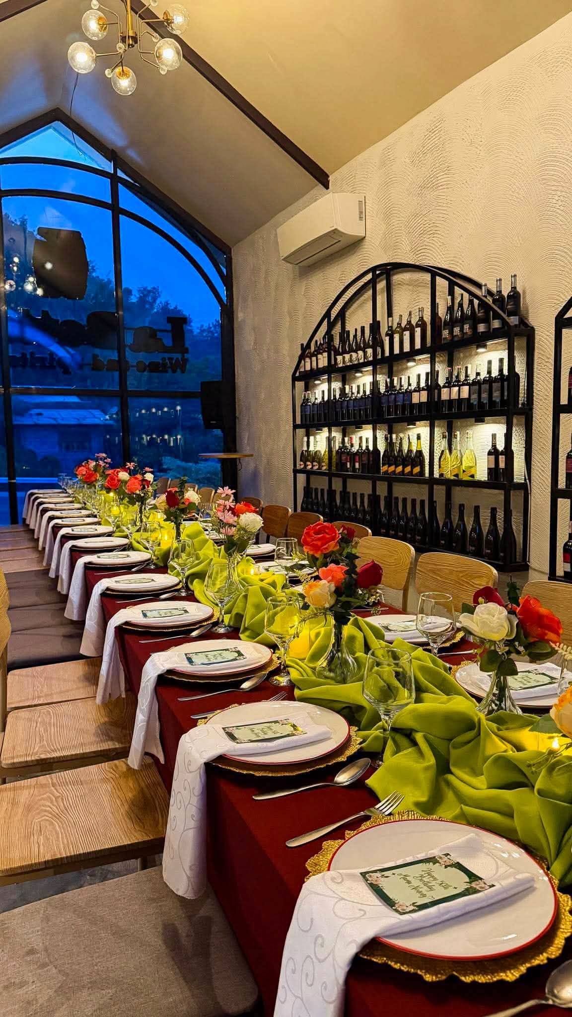 A long table dressed in red and green beneath a tall arched window and the wine wall